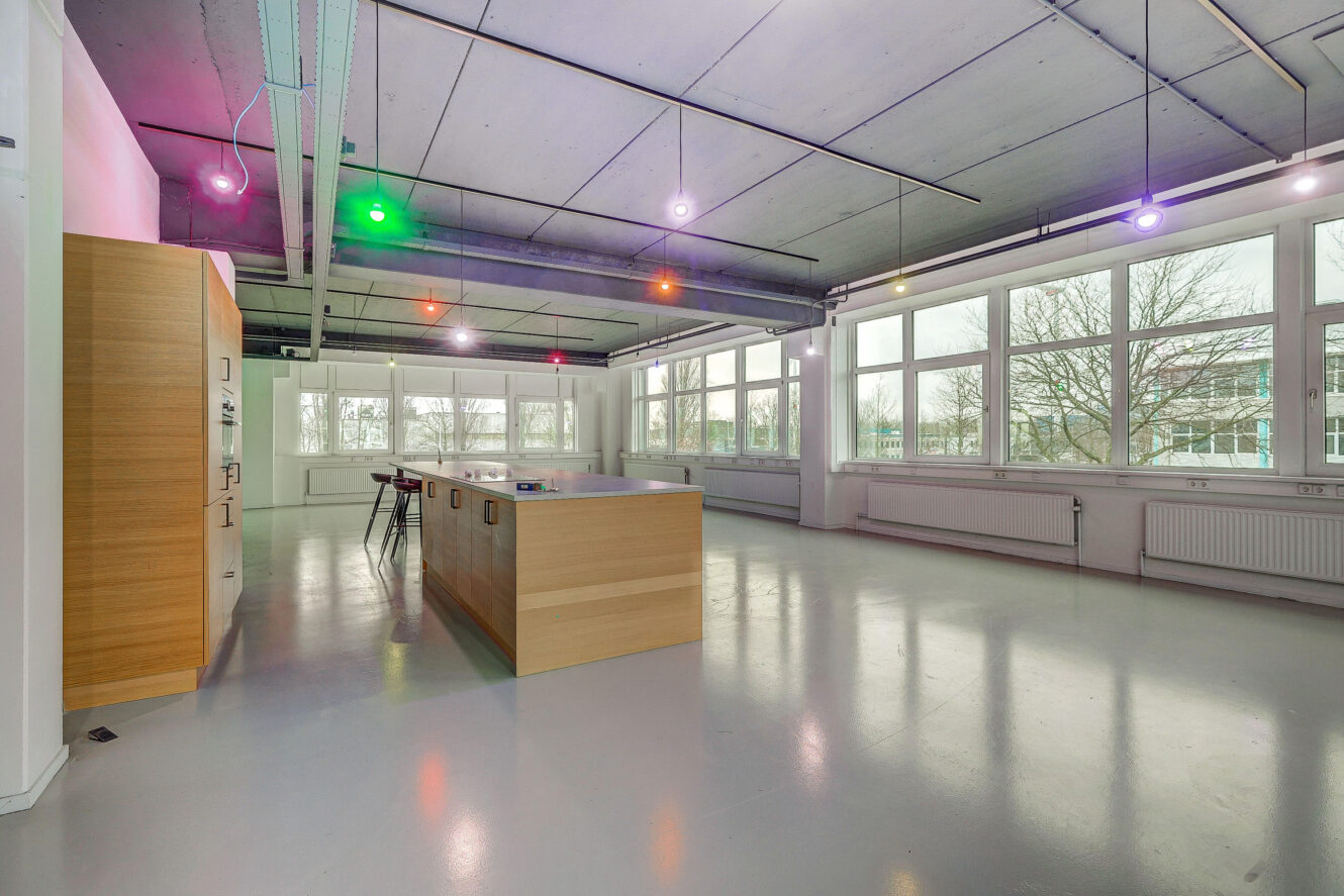 Ruime moderne kamer met grote ramen, een kookeiland, houten kasten en gekleurde plafondverlichting. De vloer is glanzend en de ruimte lijkt leeg op een paar krukjes na.