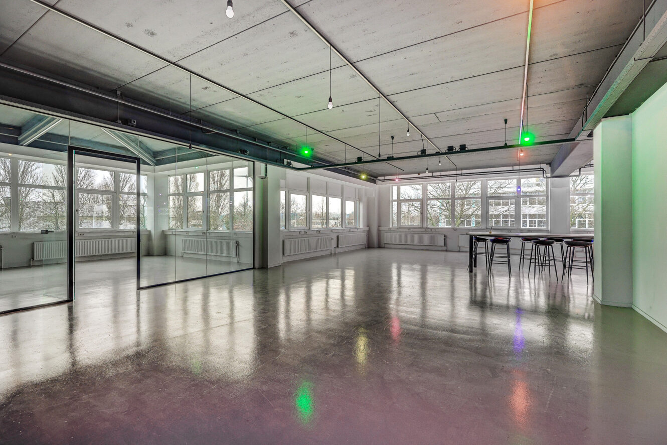Open, moderne kantoorruimte met glazen scheidingswanden, grote ramen, gepolijste betonnen vloeren en zichtbare plafondarmaturen. Tafels en krukken staan in een hoek.