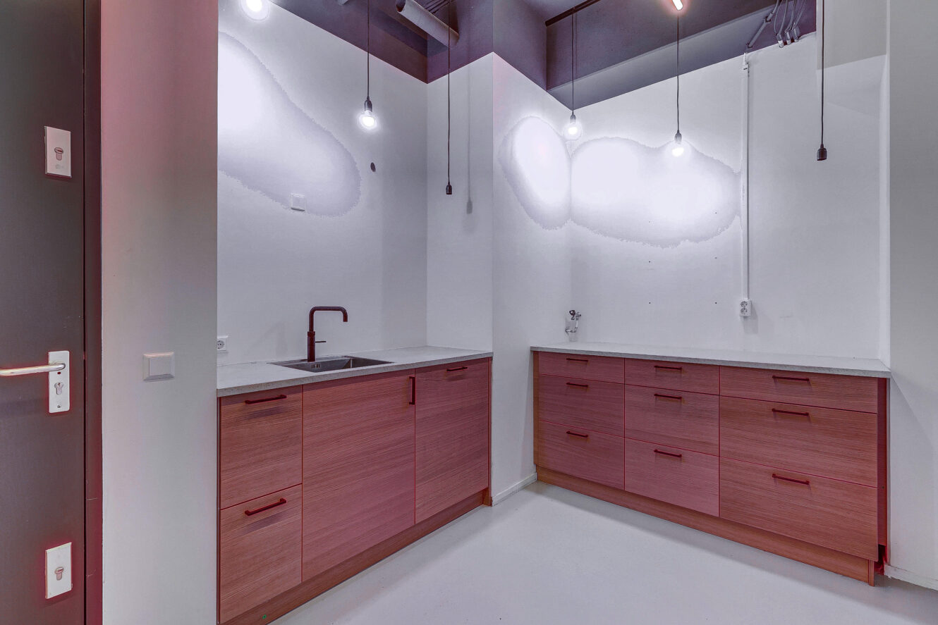 Minimalistische keuken met houten kastjes, witte werkbladen, witte muren, een gootsteen met een zwarte kraan en hangende hanglampen. Geen zichtbare apparatuur of decor.