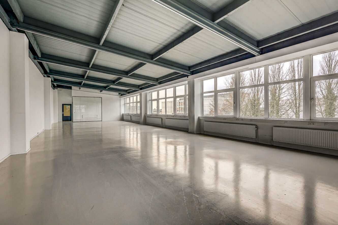 Grote, lege ruimte in industriële stijl met gepolijste betonnen vloer, balkenplafond en een muur van ramen die natuurlijk licht binnenlaten.