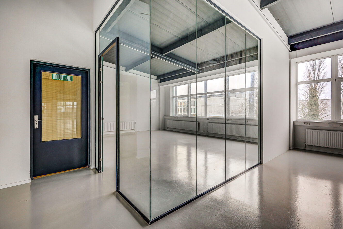 Een moderne, lege kamer met grote ramen en een kantoorruimte met glazen wanden; aan de linkermuur is een deur met het opschrift KNOOPKAMER zichtbaar.