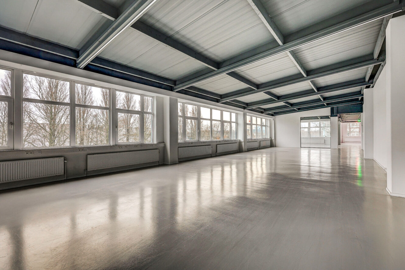 Grote, lege ruimte met gepolijste betonnen vloer, witte muren, zichtbare stalen balken en grote ramen die natuurlijk licht binnenlaten.