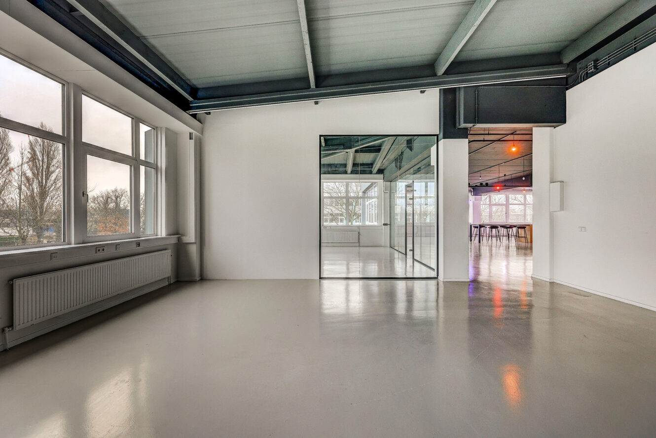 Een moderne, lege kamer met grote ramen, een gepolijste vloer, een wandspiegel en uitzicht op een helder verlichte aangrenzende ruimte.