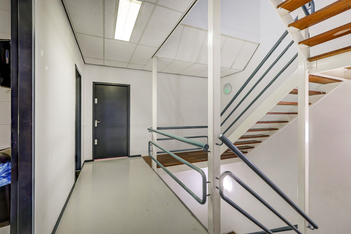 Licht trappenhuis in een modern gebouw met witte muren, houten treden, metalen leuningen en een gesloten zwarte deur op de overloop.