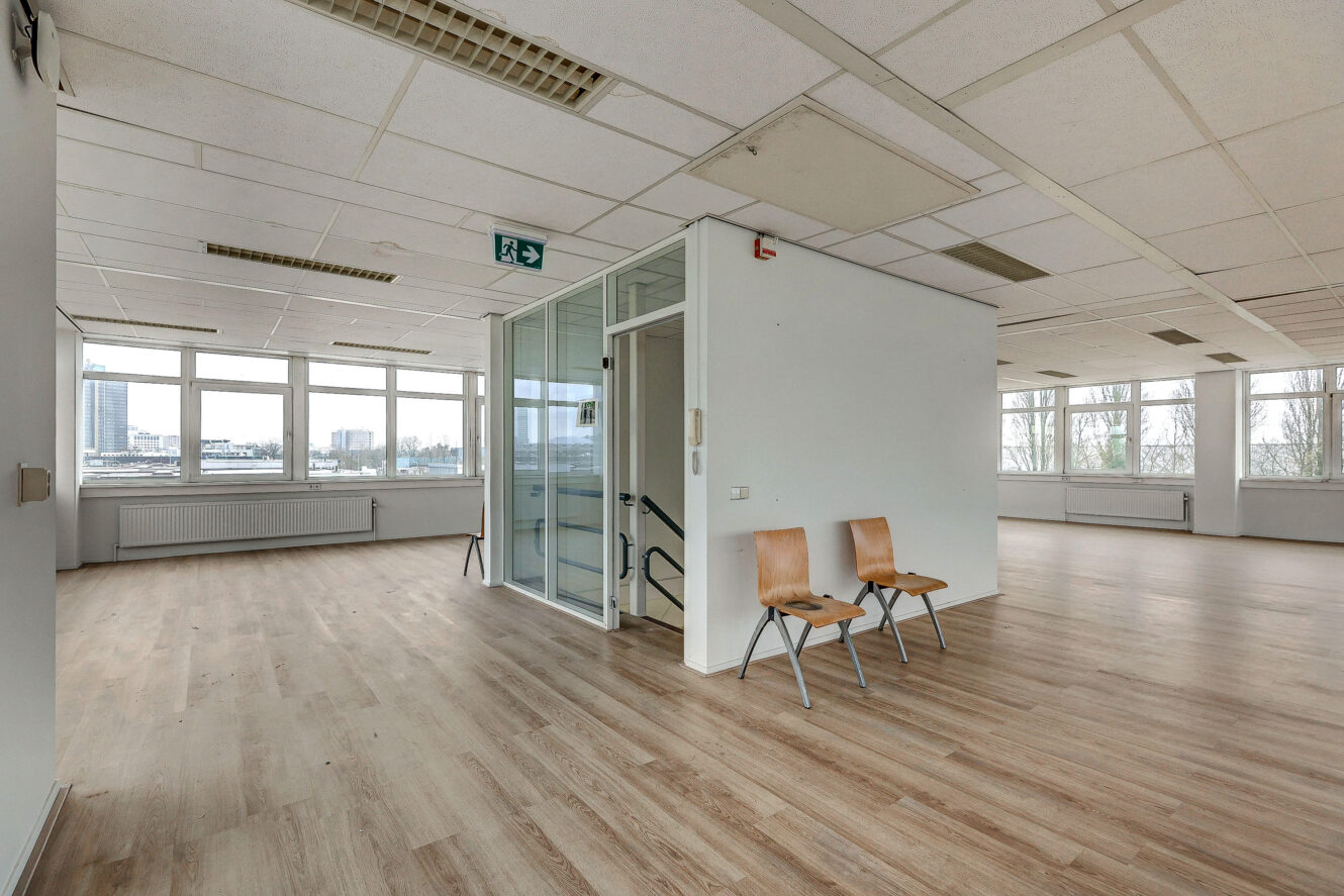 Lege kantoorruimte met houten vloeren, grote ramen, een lift met glazen panelen en twee houten stoelen tegen een witte muur.