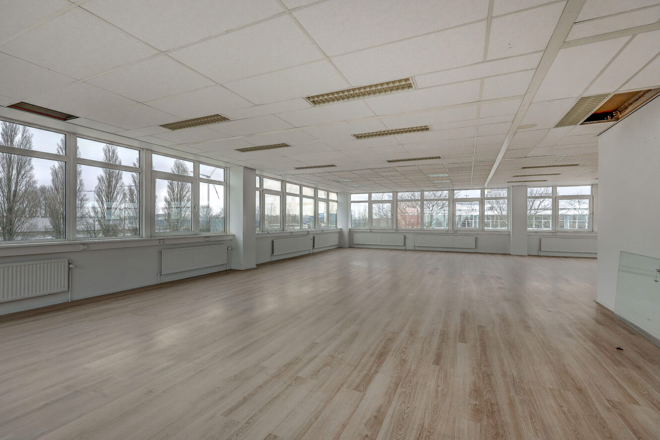 Ruime, lege kamer met grote ramen, lichte houten vloeren en een wit verlaagd plafond; sommige plafondpanelen en verlichtingsarmaturen ontbreken of zijn beschadigd.