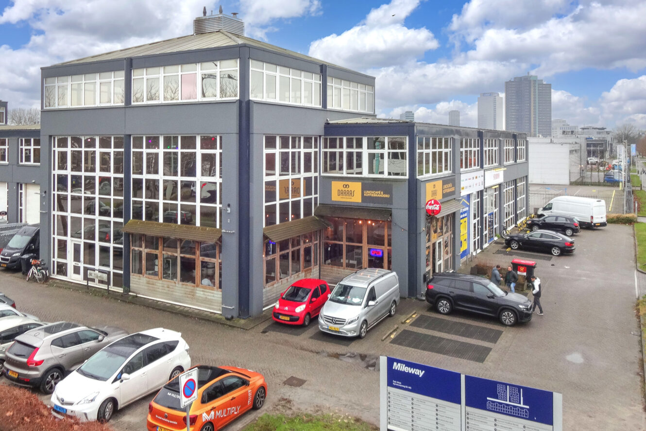 Groot commercieel gebouw van twee verdiepingen met glazen gevel, meerdere uithangborden en geparkeerde auto's aan de voorkant onder een gedeeltelijk bewolkte hemel.