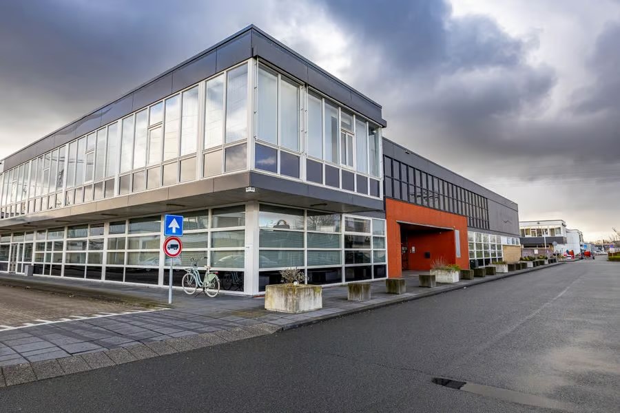 Modern kantoorgebouw van twee verdiepingen met grote ramen onder een bewolkte hemel; een fiets staat buiten geparkeerd bij een eenrichtingswegwijzer.