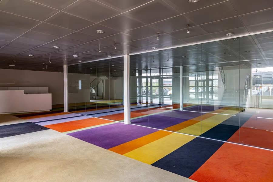 Moderne kantoorruimte met glazen muren, grijs plafond en een vloer met gedurfde strepen in oranje, geel, rood en paars; zonovergoten en leeg.