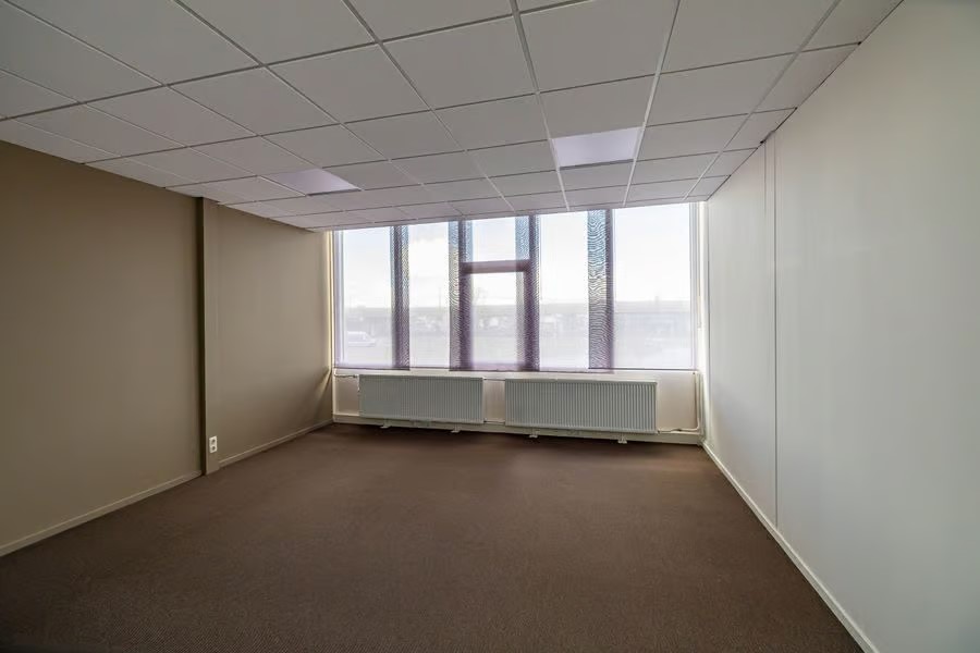 Lege kantoorkamer met bruin tapijt, witte plafondtegels, beige en witte muren, een groot raam en twee radiatoren eronder.