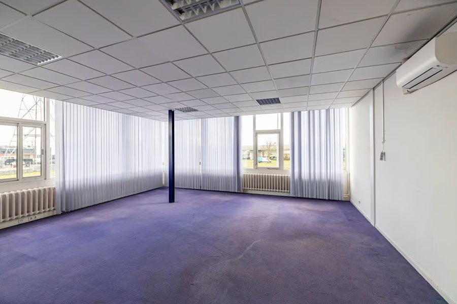 Een leeg kantoor met paars tapijt, witte muren, grote ramen met verticale jaloezieën, bovenlichten en een centrale pilaar.