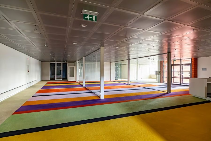 Een ruime kamer met kleurrijk gestreept tapijt, kantoren met glazen wanden, witte muren en een grijs betegeld plafond met een bordje met de uitgang.