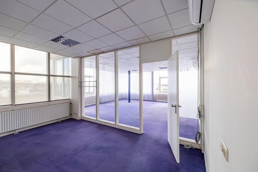 Leeg kantoor met paars tapijt, grote ramen, glazen scheidingswanden en een open deur naar een andere kamer. Plafondtegels en lampen boven het hoofd.