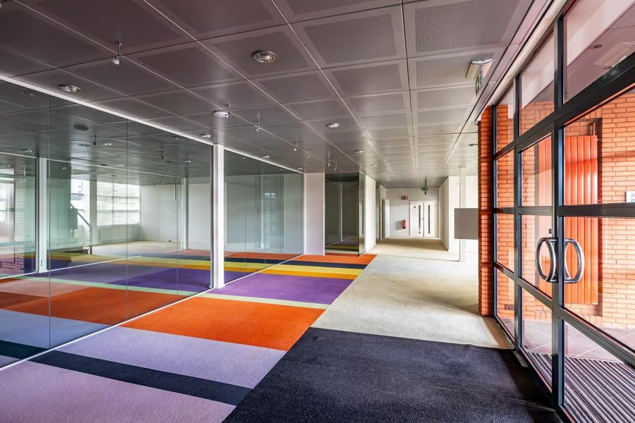 Moderne kantoorruimte met glazen muren, levendige geometrische tapijten, zichtbare plafondpanelen en grote glazen toegangsdeuren.