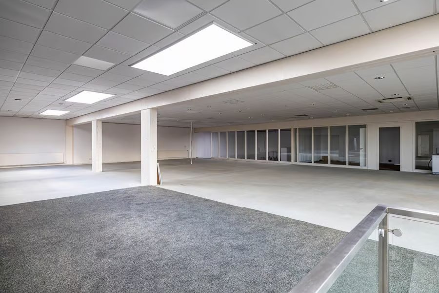 Ruime lege kantoorkamer met witte muren, plafondverlichting, grote ramen en grijs tapijt op de voorgrond.