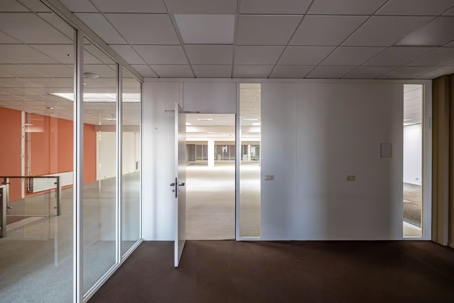Lege kantoorruimte met glazen muren, een open deur, witte plafondtegels en zowel vloerbedekking als kale vloeren onder tl-verlichting.