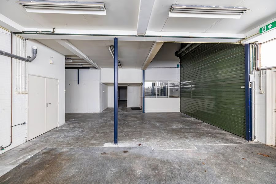Interieur van een leeg industrieel magazijn met betonnen vloer, witte muren, groene roldeur, TL-verlichting en pijpen.