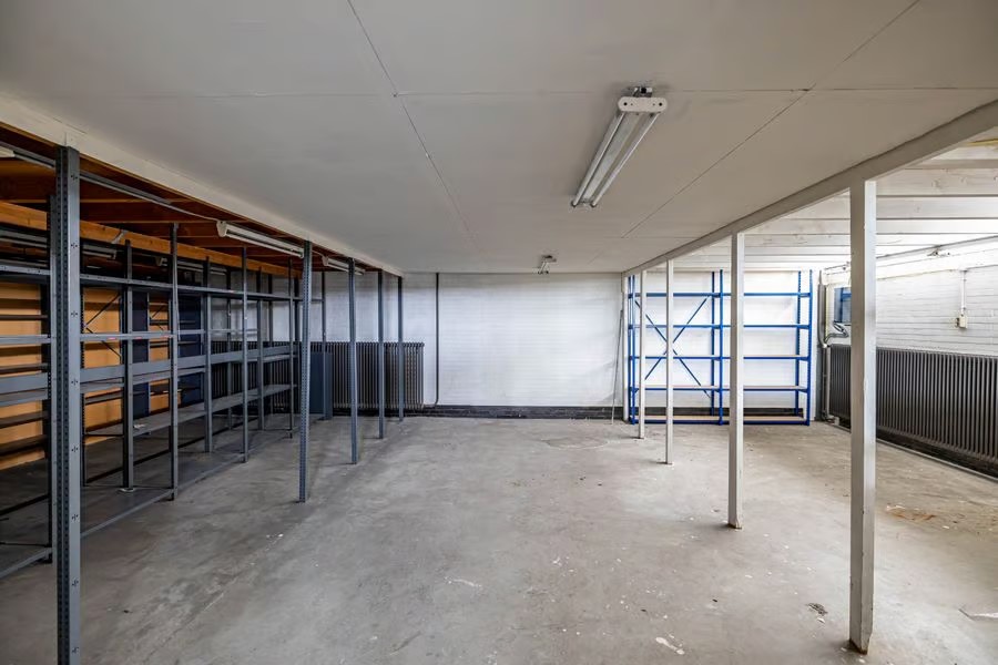 De lege industriële ruimte heeft een betonnen vloer, metalen rekken langs de muren, tl-verlichting en wit geschilderde oppervlakken.