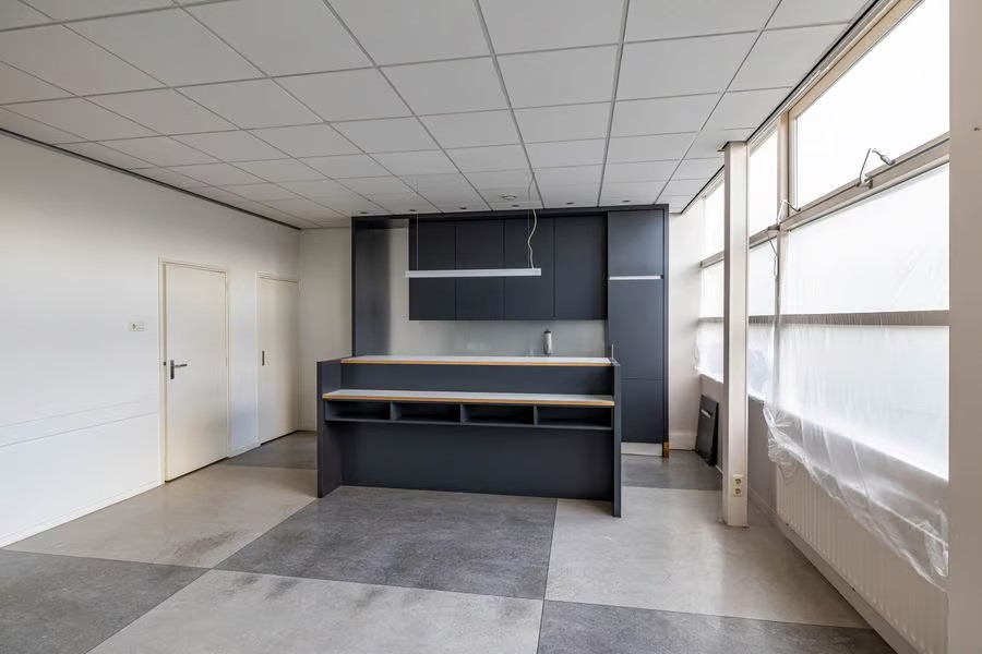 Moderne, minimale keuken met donkere kasten, centraal eiland, grote ramen en betegeld plafond in een lichte, ongemeubileerde kamer.