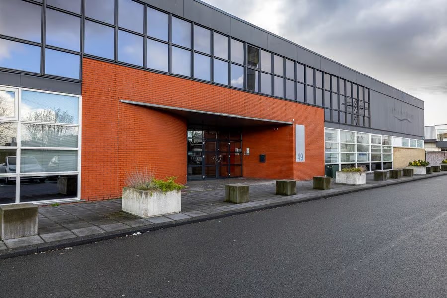 Modern commercieel gebouw met grote glazen ramen, rode bakstenen gevel en betonnen plantenbakken langs een geplaveid trottoir onder een bewolkte hemel.