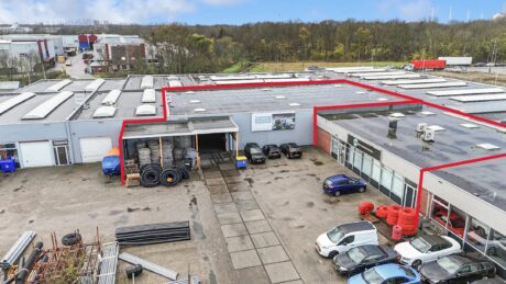 Industrieel magazijncomplex met geparkeerde voertuigen, opgerolde pijpen en opslagmateriaal buiten; gebouw rood omlijnd. Bomen erachter.