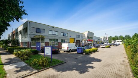 Industriegebouw met meerdere units, auto’s en bestelwagens die op een geasfalteerde weg geparkeerd staan, groen aan beide kanten, een helderblauwe lucht erboven.