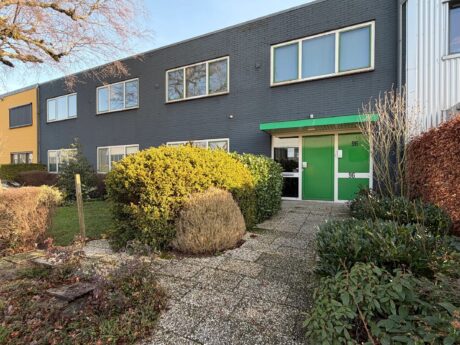 Een gebouw van twee verdiepingen met donkergrijze bakstenen muren, grote ramen en een groen-witte deur. Een pad loopt door de struiken naar de ingang.