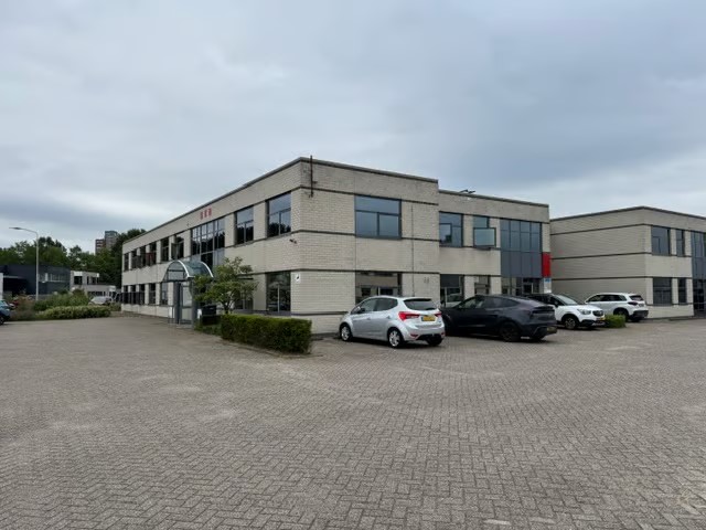 Een kantoorgebouw van twee verdiepingen met grote ramen en een aantal geparkeerde auto’s ervoor, onder een bewolkte hemel.