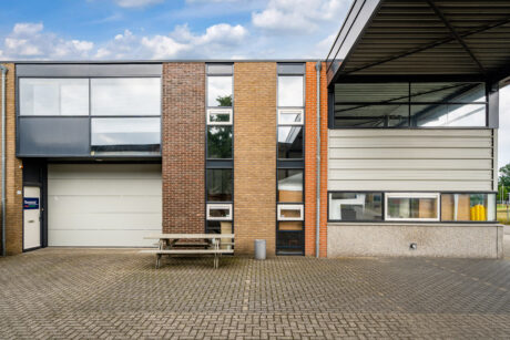 De buitenkant van een modern industrieel gebouw met een gevel die bestaat uit een combinatie van baksteen en metaal, grote ramen, een garagedeur, een picknicktafel en een vuilnisbak, gelegen op een bestraat terrein onder een gedeeltelijk bewolkte hemel.
