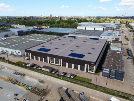 Luchtfoto van een groot pakhuis met zonnepanelen op het dak, omringd door industriële gebouwen en geparkeerde voertuigen onder een gedeeltelijk bewolkte hemel.