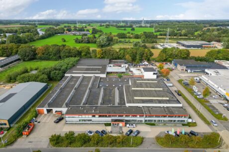 Luchtfoto van een groot complex van bedrijfsgebouwen met een plat dak, omgeven door parkeerterreinen en groene velden.