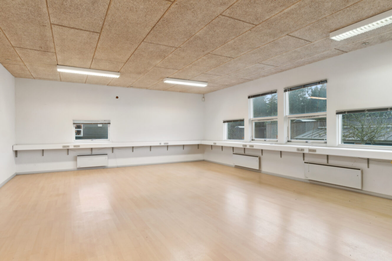 Tomt rum med lyst trægulv, hvide vægge, flere vinduer, lysstofrør i loftet og vægmonterede bordplader langs siderne.