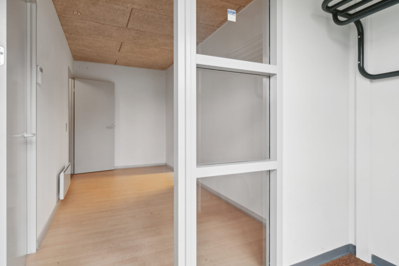 Et lille, tomt rum med lyst trægulv, hvide vægge, en glasvæg og en dør, der fører til et andet rum.