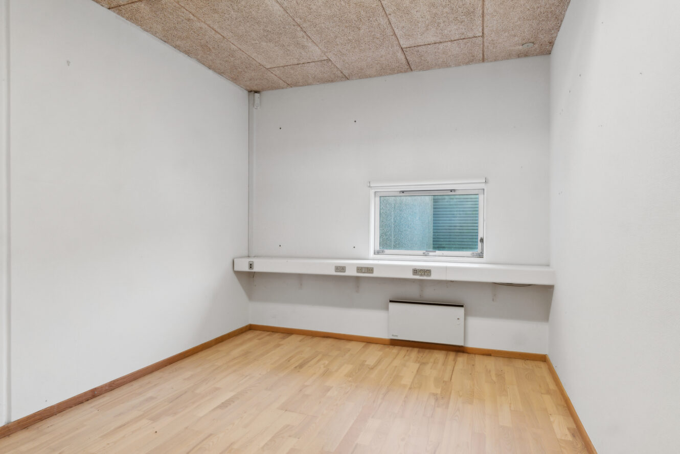 Tomt rum med hvide vægge, et lille vindue, indbygget hvidt skrivebord, trægulv og et loft med akustikplader.