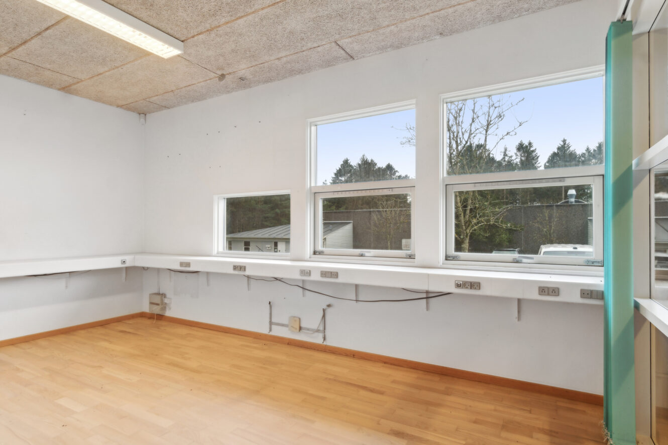 Tomt rum med lyst trægulv, hvide vægge, store vinduer, stikkontakter langs væggen og synlige kabler under en hylde i bordhøjde.