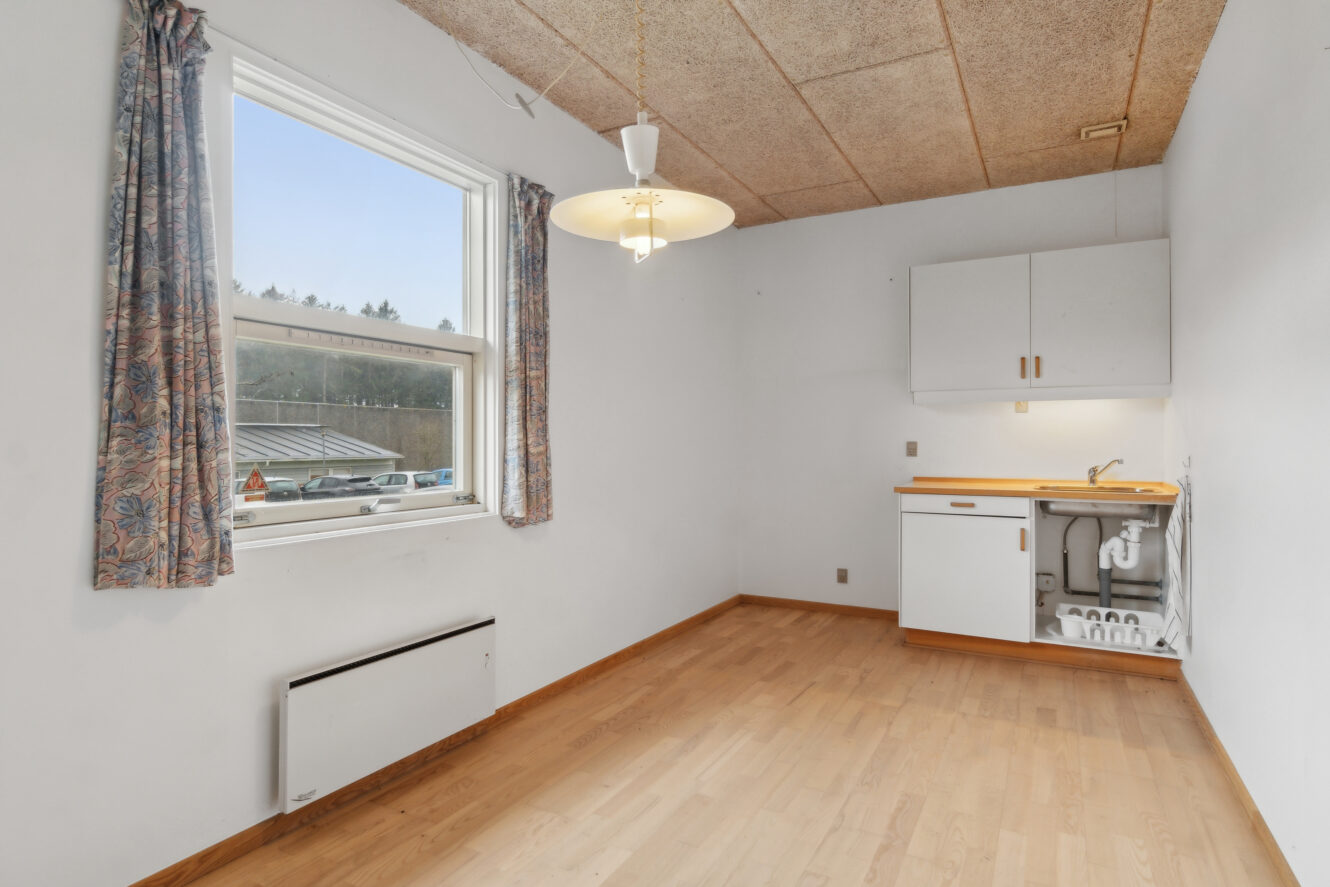 Lille rum med lyst trægulv, et vindue med mønstrede gardiner, en loftslampe, en vægvarmer og et tekøkken med en synlig vask og et skab.