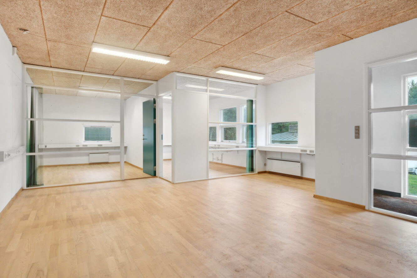 Tomme, moderne kontorlokaler med lyse trægulve, hvide vægge, glasskillevægge, loftslamper og flere vinduer, der giver naturligt lys.