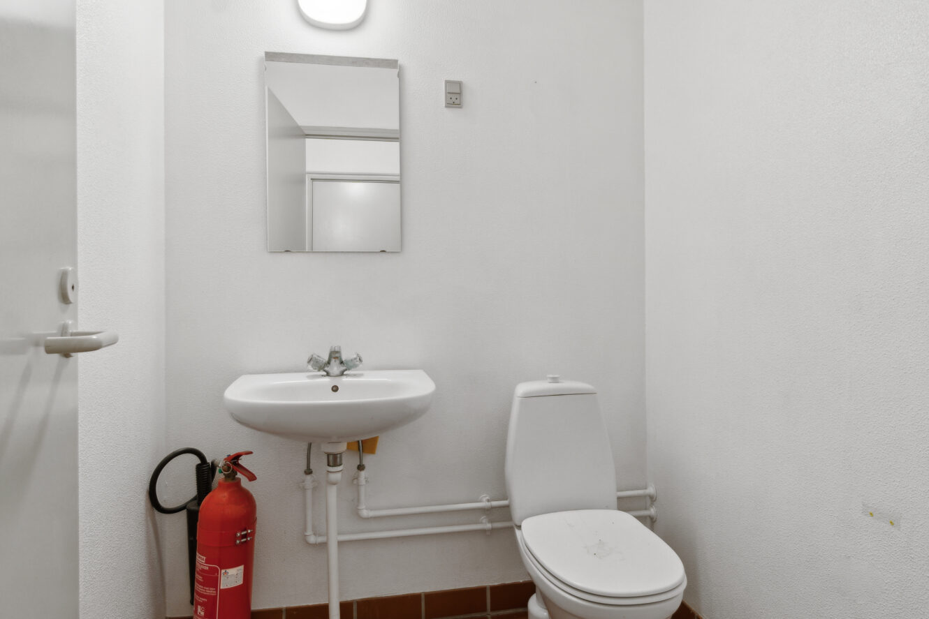 Lille badeværelse med hvidt toilet, væghængt håndvask, spejl og en rød brandslukker placeret på gulvet ved siden af håndvasken.