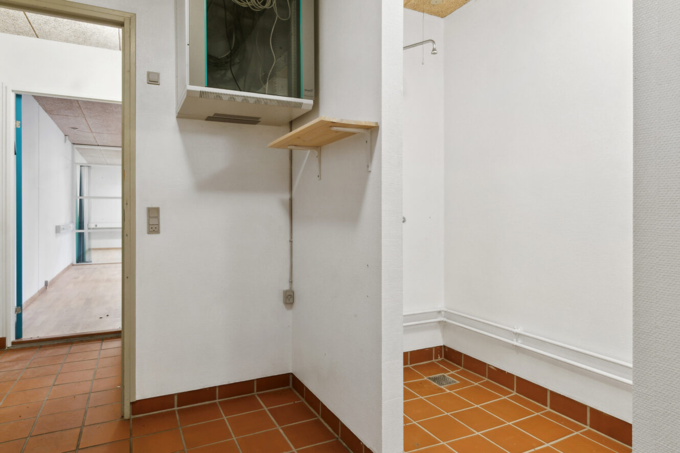 Lille bryggers med hvide vægge, rødt flisegulv, synlige ledninger i et overliggende panel, en lille træhylde og delvis udsigt til et tilstødende rum med trægulv.