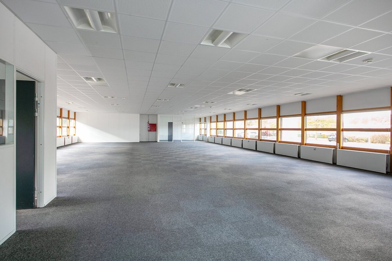 Stort, tomt kontorlokale med gulvtæppe, hvide vægge og rækker af vinduer, der lukker naturligt lys ind langs højre side.