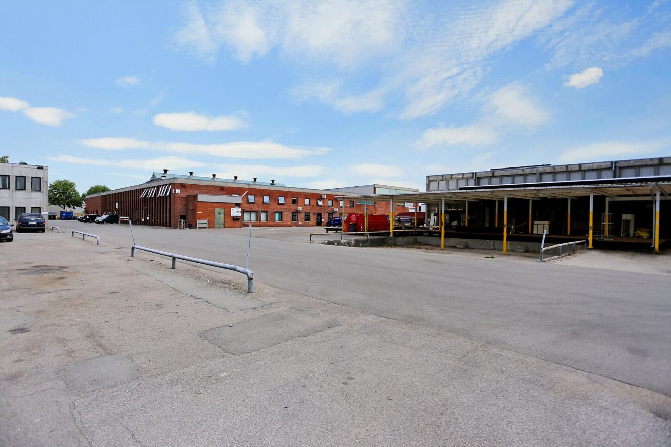 Stor industriparkeringsplads med få parkerede biler, en rød murstensbygning til venstre og et metaloverdækket læsseområde til højre under en delvis overskyet himmel.