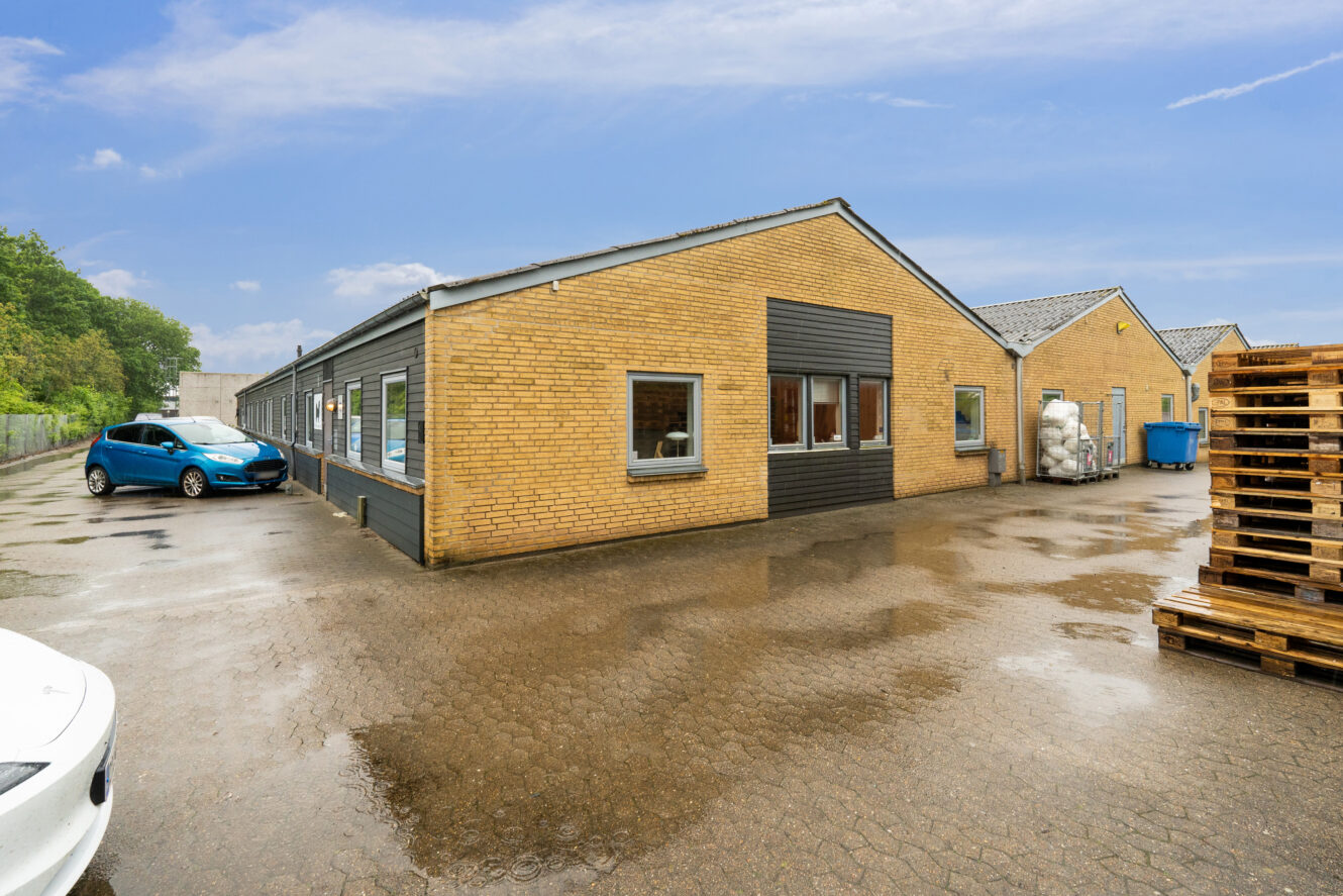 Industriel murstensbygning i én etage med store vinduer, parkerede biler, træpaller og blå containere på en våd asfalteret plads under en delvis overskyet himmel.