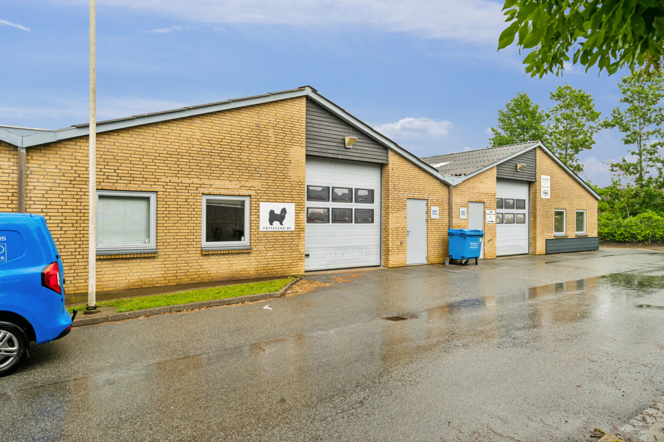 Gul murstensindustribygning i én etage med to garageporte, flere vinduer, en blå varevogn, en blå skraldespand og et vådt fortov, der afspejler den seneste tids regn.
