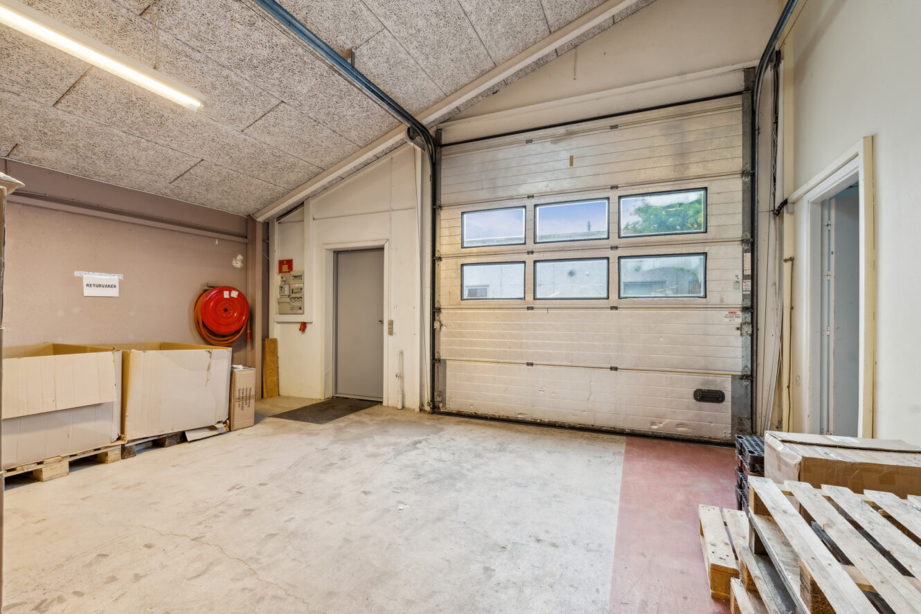 Et lagerinteriør med en lukket garageport, stablede papkasser, træpaller, en brandslangetromle og en grå dør på venstre væg.