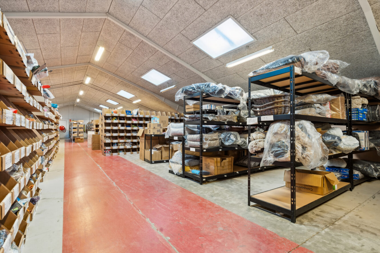 Lagerinteriør med hylder med forskellige varer, nogle pakket ind i plastik, og kasser organiseret langs en gang under kraftig ovenlys.