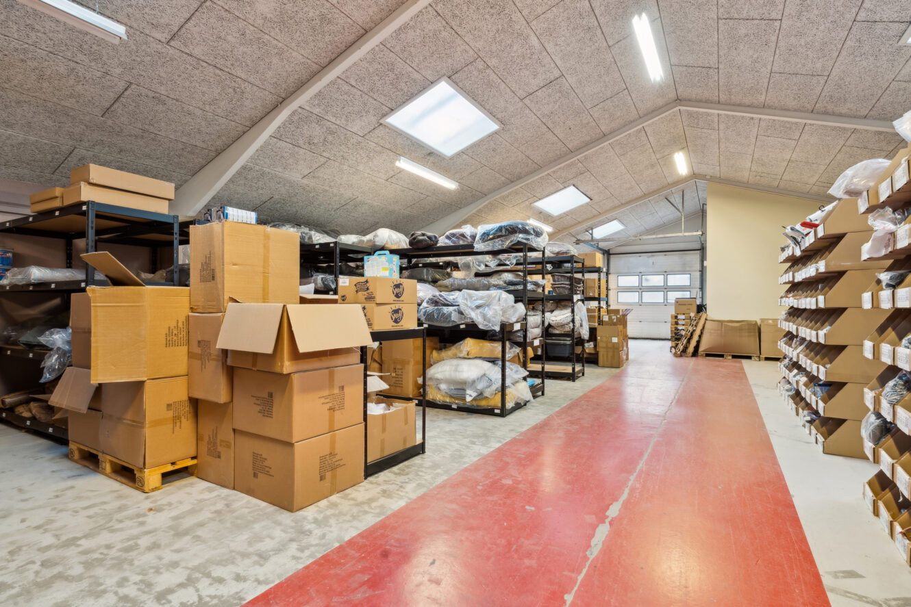 Lageret er indrettet med hylder og stativer med kasser og poser, nogle kasser er åbne og stablet på gulvet, og en rød gangbro løber ned gennem midten af lokalet.