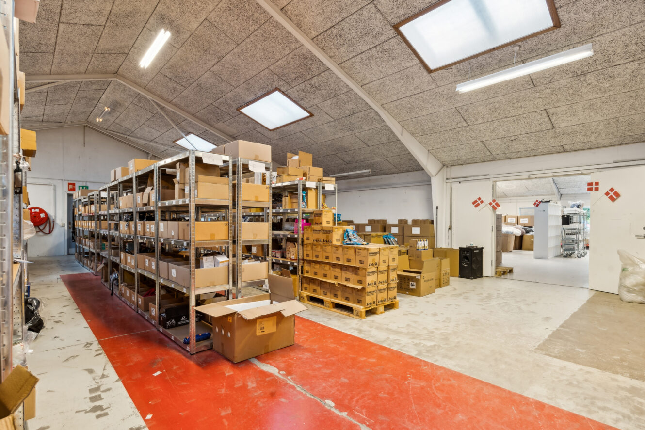Industrielt lagerinteriør med metalreoler fyldt med kasser og pakker, rødt og hvidt gulv og lysstofrør over hovedet.