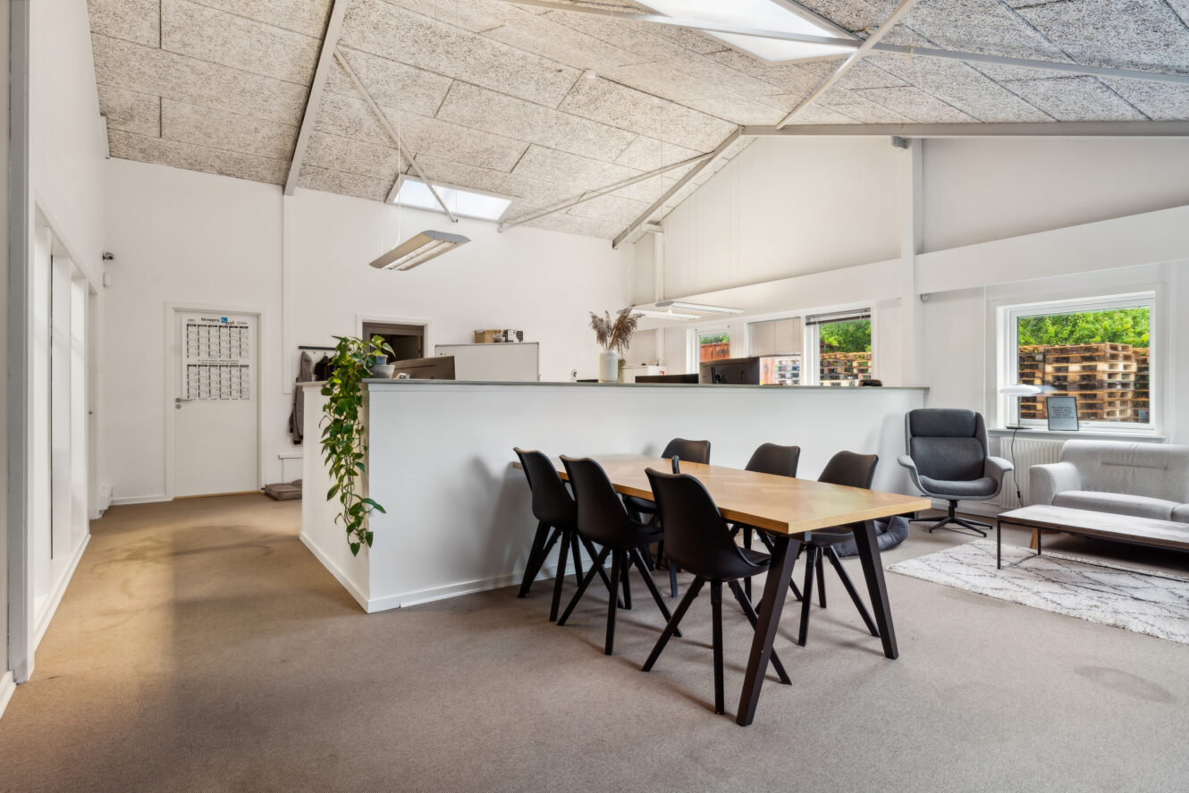 Moderne kontorlokaler med mødebord i træ og sorte stole, hvide vægge, gulvtæppe, loungeområde, store vinduer og naturligt lys.