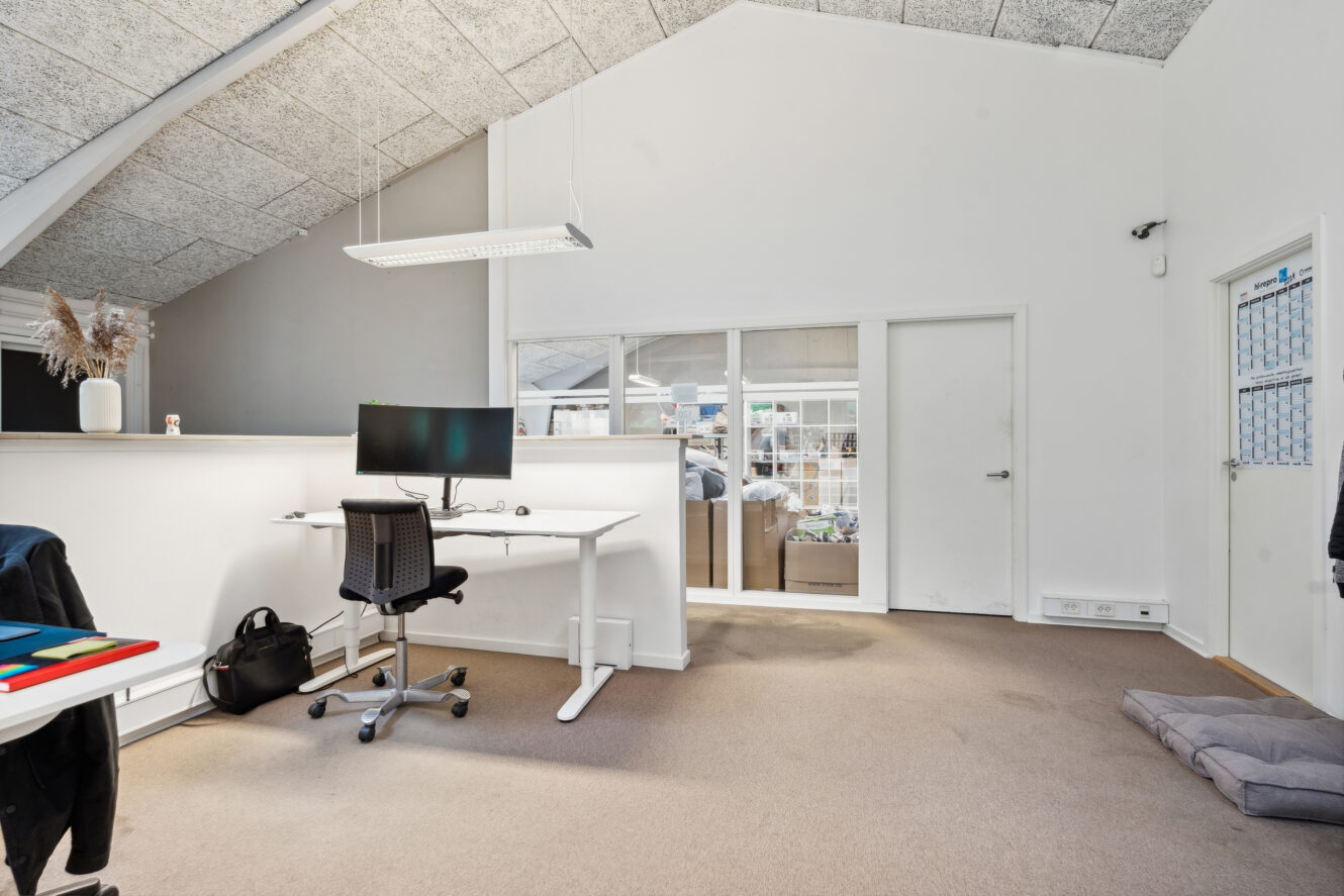 Moderne kontor med et hvidt skrivebord med en computerskærm, en sort stol, en taske på gulvet og en glasvæg, der afslører et opbevaringsområde bagved.