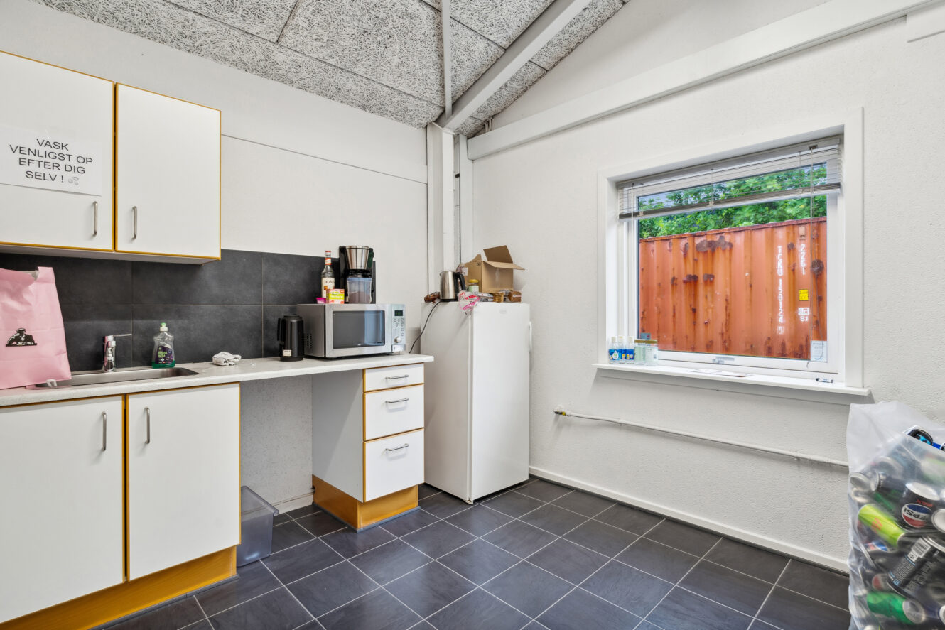 Et lille tekøkken med køleskab, mikroovn, kaffemaskiner og skabe; et vindue viser en skibscontainer udenfor.